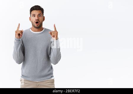 Surprised hispanic bearded man with stylish modern hairstyle, open mouth wondered, gasping fascinated with admiration, pointing up impressed with Stock Photo