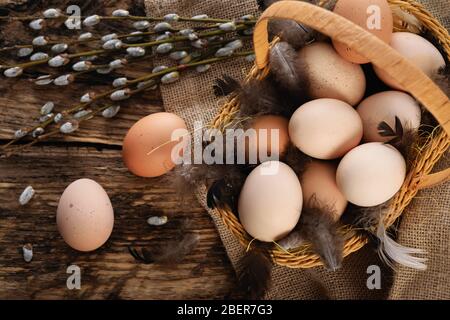 Easter composition in vintage style - wicker basket with eggs, feathers and willow cats on a burlap sack and a wooden table Stock Photo