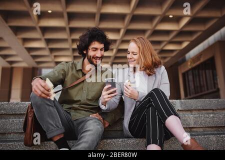 Cheerful woman in formal clothing showing man video on smartphone while eating sandwich during break sitting on stairs Stock Photo