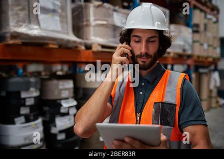 Focused warehouse manager tracking supply order details using digital tablet while in conversation over mobile phone in warehouse Stock Photo