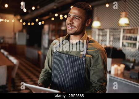 Handsome young man wearing apron holding digital tablet standing in coffee house looking away