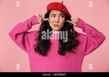 Image of upset beautiful asian girl with long dark hair wearing beret plugging her ears with fingers isolated over pink background Stock Photo