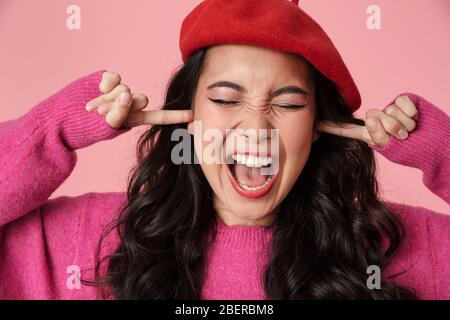 Image of upset beautiful asian girl wearing beret screaming and plugging her ears with fingers isolated over pink background Stock Photo