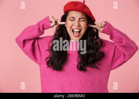 Image of upset beautiful asian girl wearing beret screaming and plugging her ears with fingers isolated over pink background Stock Photo
