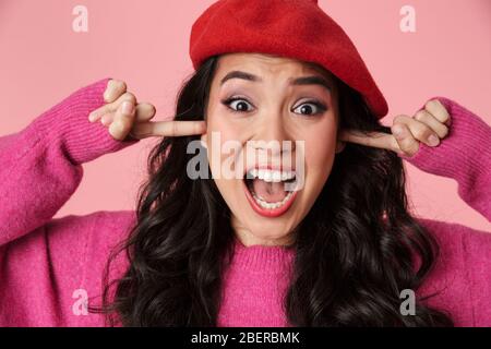 Image of upset beautiful asian girl wearing beret screaming and plugging her ears with fingers isolated over pink background Stock Photo