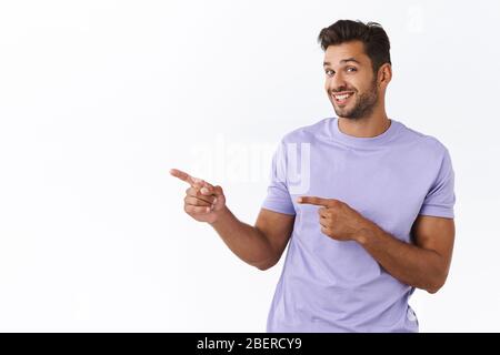 Silly smiling gay man invite check out and see nice offer, pointing left and gazing camera with satisfied outgoing, friendly grin, recommend promo Stock Photo