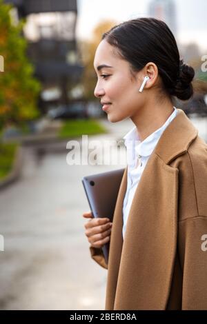 Attractive young asian businessowoman wearing coat walking outdoors in the city, carrying laptop Stock Photo