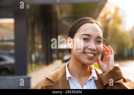 Image of young brunette asian woman wearing coat using earbuds while walking through city street Stock Photo