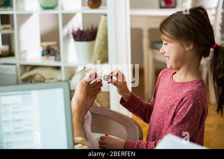 Child playing and disturbing father working remotely from home. Little girl applying nail polish on toenails. Man sitting on couch with laptop. Family Stock Photo
