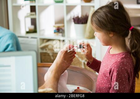 Child playing and disturbing father working remotely from home. Little girl applying nail polish on toenails. Man sitting on couch with laptop. Family Stock Photo