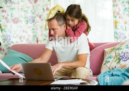 Child playing and disturbing father working remotely from home. Man sitting on couch with laptop. Family spending time together indoors. Stock Photo