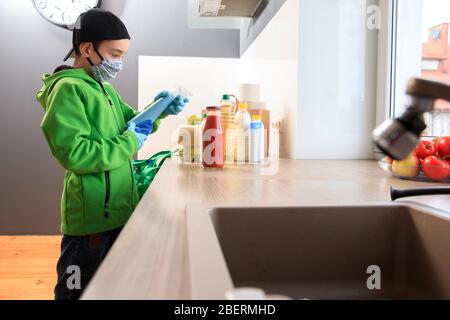 Coronavirus pandemic prevention. A boy wearing gloves and a mask uses a disinfectant liquid to wipe the food package upon delivery at home. Cleaning t Stock Photo