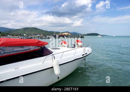 Two men in white and red uniform clean a white yacht moored at the dock of Koh Samui in Thailand. Beautiful view of the wooden pier and mountains Stock Photo