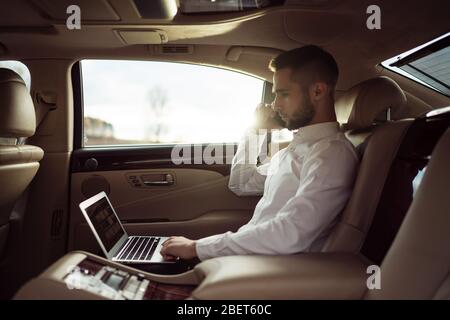Man uses laptop and talking on smartphone sitting in back seat of car, businessman in taxi Stock Photo