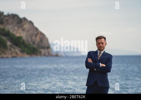 Businessman standing on the beach in a suit. Looks at camera and smiles Stock Photo