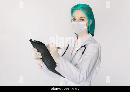 Portrait of young pretty female doctor wearing white coat, medical cap and face mask, holding clipboard and writing notes, looking at camera, standing Stock Photo