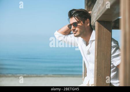 Young attractive man with sunglasses looking out over the sea during the summer. He looking forward, dressed in a white shirt and leaning on a wooden Stock Photo