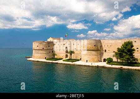 Old medieval Aragonese Castle with palm trees in front of blue sky with white clouds on sea channel in historic center of Taranto city, Puglia (Apulia Stock Photo