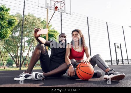 Outdoors Activity. African couple sitting on basketball court taking selfie on smartphone smiling joyful Stock Photo