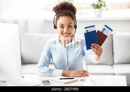 Tour agent in headset sitting at table in office holding passports and tickets looking camera cheerful Stock Photo