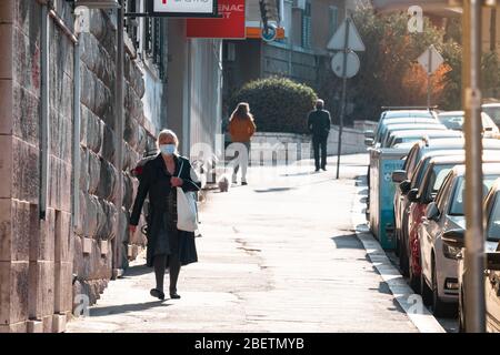 Older woman in a black coat and a white surgical face mask walking down the street of Split during coronavirus outbreak pandemic quarantine time Stock Photo