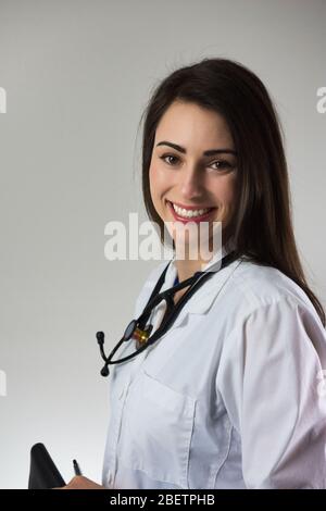 Female medical professional holding stethoscope around neck isolated on grey background. Attractive young female with beautiful smile. Stock Photo