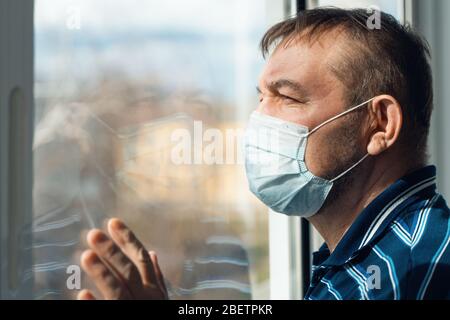 Portrait of an elderly man in a medical mask. Senior man at home looks out of the window during quarantine. Coronavirus, COVID-19 outbreak. Stock Photo