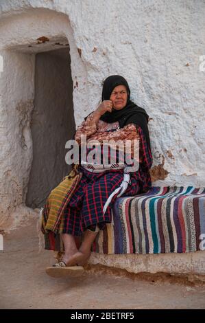 Old berber woman sitting on a bench covered with colorful rug in front of her traditional white house in Tunisia Stock Photo