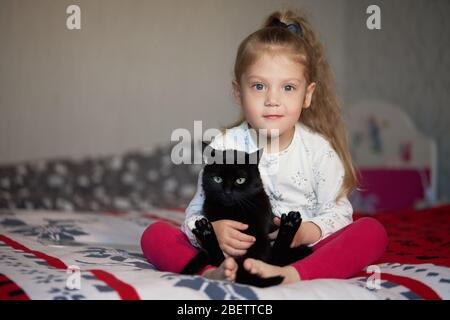Portrait of a little cute child girl who hugs a black cat with tenderness and love and smiles with happiness Stock Photo