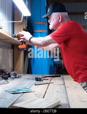 Senior man putting up a shelf in his shop. He is drilling the screws into the wall Stock Photo