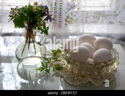 Forest primroses in a retro glass bottle and a crystal vase with eggs on a glass surface . Stock Photo