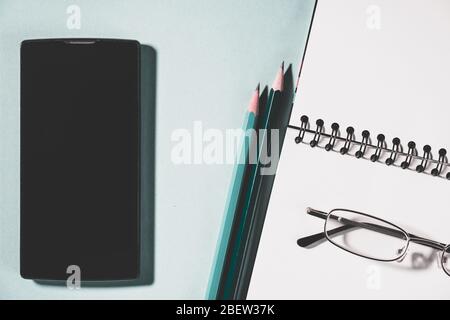 top view of cell phone, pencils and school notebook on green background, glasses and spiral notepad on a table. High angle view of empty planner. Stock Photo