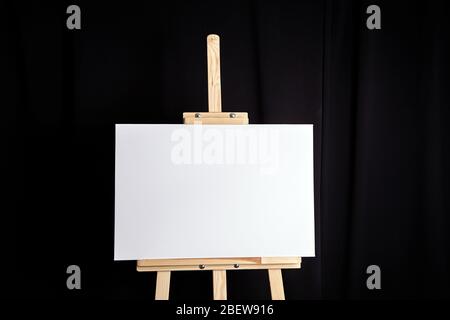 White blank canvas stands on a wooden artistic easel on black