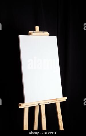 White blank canvas stands on a wooden artistic easel on black