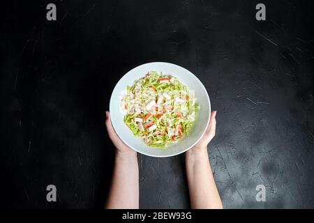 Chopped crab sticks and fresh chinese cabbage leaves in white ceramic bowl on black stone background, top view. Vegetable and imitation crab meat, sea Stock Photo