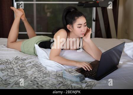 Portrait attractive young Asia woman Lie down on the bed working on a laptop in the bedroom with stack 100 US dollars banknotes a lot of, works from o Stock Photo