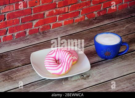 Two fresh pink glazed donuts and a blue cup of cappuccino stand on a wooden table against a red brick wall. Close-up. Stock Photo