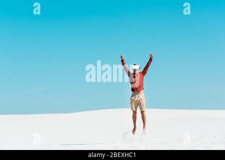 man on sandy beach in vr headset jumping against clear blue sky Stock Photo