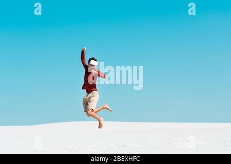 man on sandy beach in vr headset jumping against clear blue sky Stock Photo