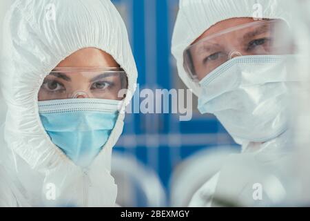 selective focus of biochemists in hazmat suites, medical masks and goggles looking at camera Stock Photo