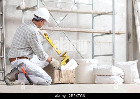 plasterer man construction worker takes the spirit level from the toolbox wear gloves, hard hat and protection glasses at interior building site with Stock Photo