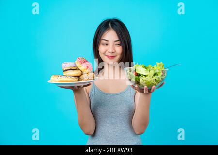 Attractive Asian young woman holding and choosing between disk of donuts or vegetable salad in glasses bowl on isolated blue color background, weight Stock Photo