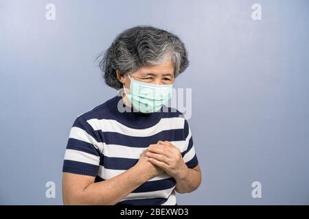 Asian Old woman wearing face surgical mask and  feeling pain ache touching chest having heart attack on blue color background, group risk of covid19 o Stock Photo