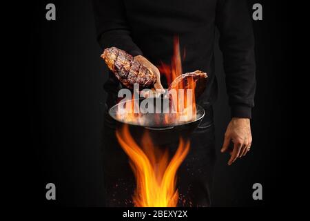 Young male dressed in jumper and jeans. He is holding burning wok pan with smoke above fire and frying two beef steaks, black background. Close up Stock Photo