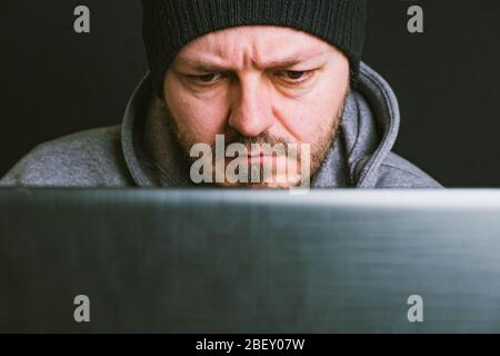 Man wearing black cap and hoodie sitting behind the computer monitor in the dark, close up, hacking computer system concept Stock Photo
