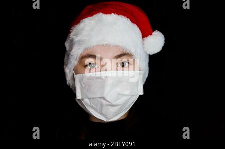 A young girl in a protective mask in a Santa Claus hat. Portrait of a brunette girl on a black background. Stock Photo