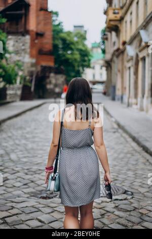 Young woman on a tour looks at beautiful homes in the city center. Girl in sundress with blue handbag and sunglasses. Girl walks the city in a dress. Stock Photo
