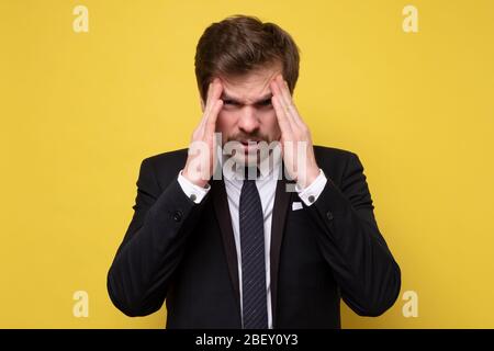 Mature businessman touching head feeling headache looking down unhappy on yellow wall. Studio shot. Negative facial human emotion. Stock Photo