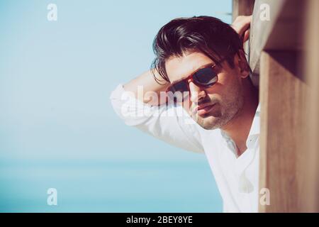 Young attractive man with sunglasses looking out over the sea during the summer. He looking forward, dressed in a white shirt and leaning on a wooden Stock Photo