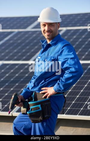 Portrait of man engineer in blue suit and protective helmet installing solar photovoltaic panel system using screwdriver. Professional electrician smiling to the camera and standing on roof of house Stock Photo